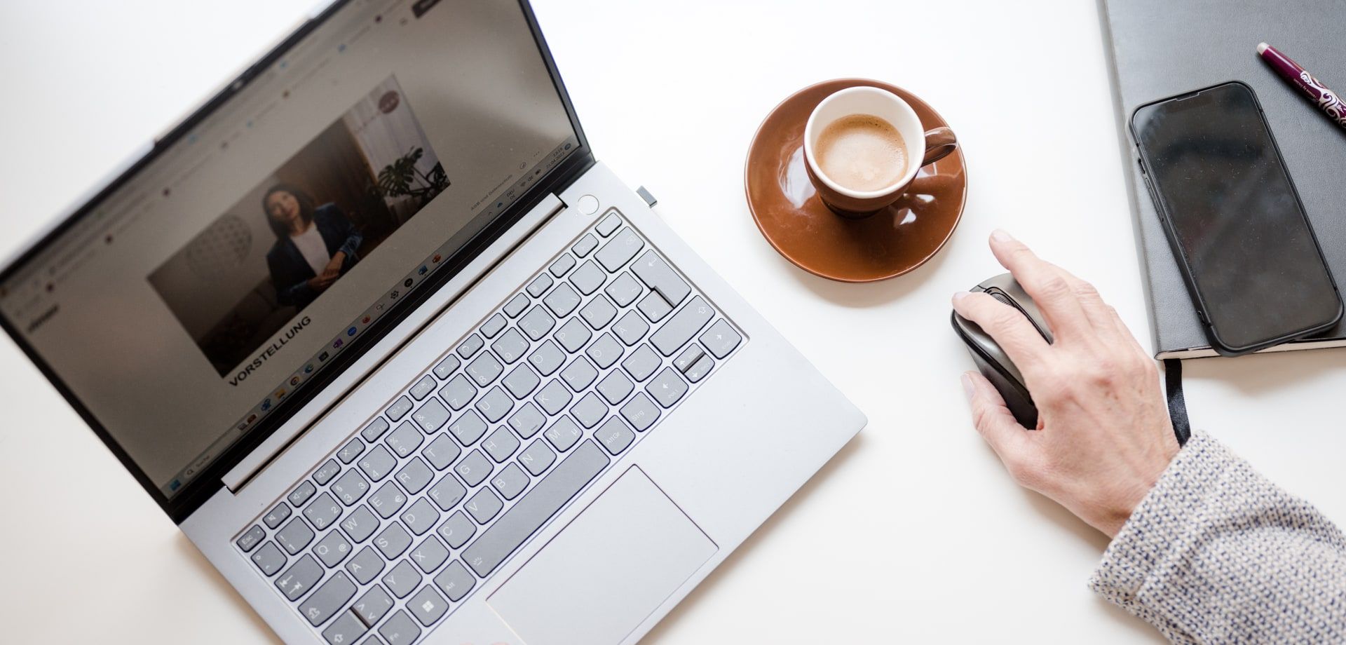 Vogelperspektive auf einen Arbeitsplatz: Laptop mit geöffneter Präsentation, eine Hand an der Maus, daneben eine Tasse Kaffee, Smartphone und Notizbuch.