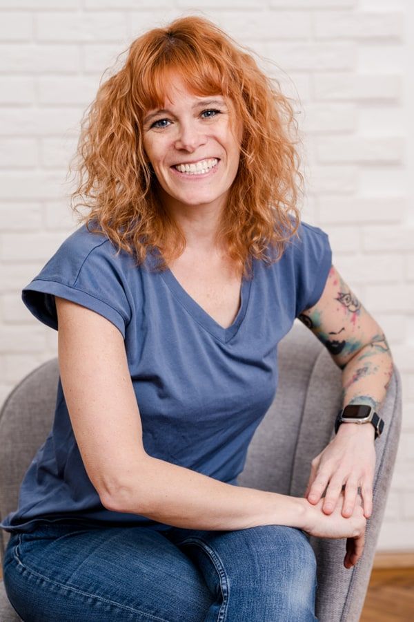 Miriam Mehlman lächelt in die Kamera wärhend sie auf einem grauen Stuhl vor weißer Ziegelwand sitzt.