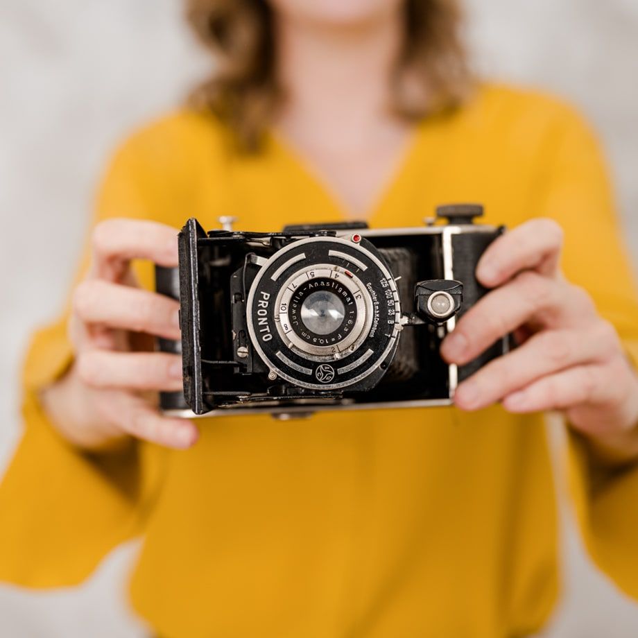 Eine Frau in gelbem Oberteil hält eine alte Faltkamera mit „Pronto"-Verschluss in die Kamera – der Fokus liegt auf der Kamera.
