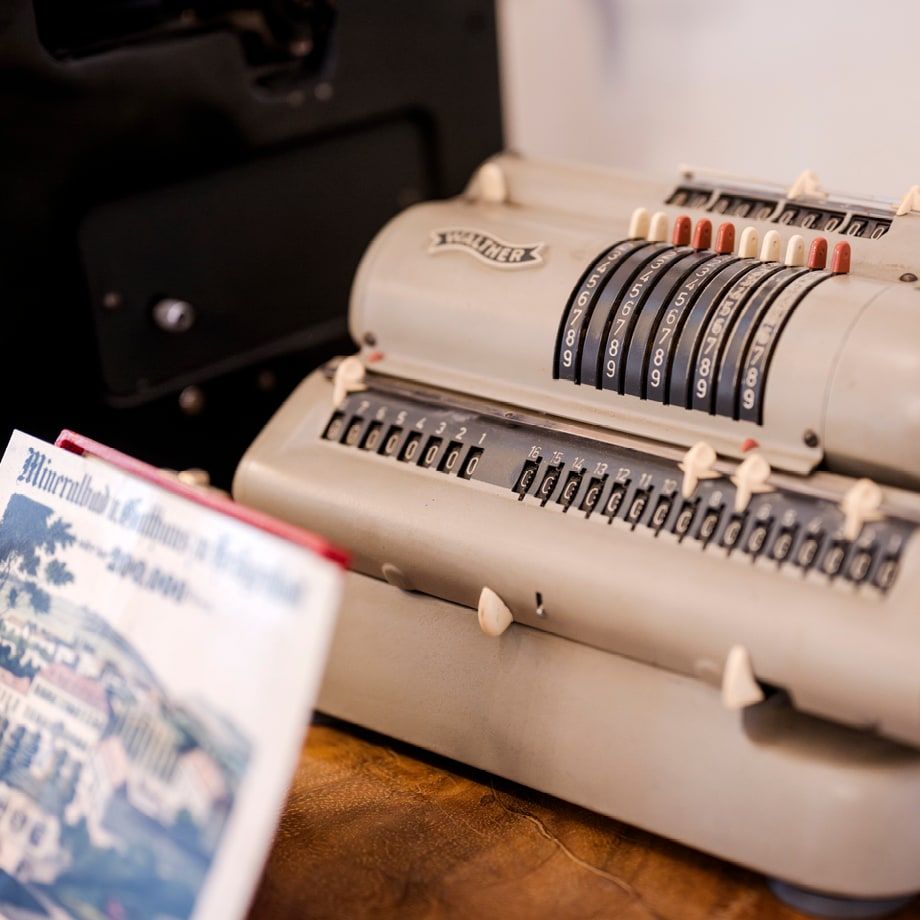 Vintage Walther-Rechenmaschine in Grau mit schwarzen Einstellrädern und Zählwerk, daneben ein altes Sparbuch – Nahaufnahme auf Holztisch.