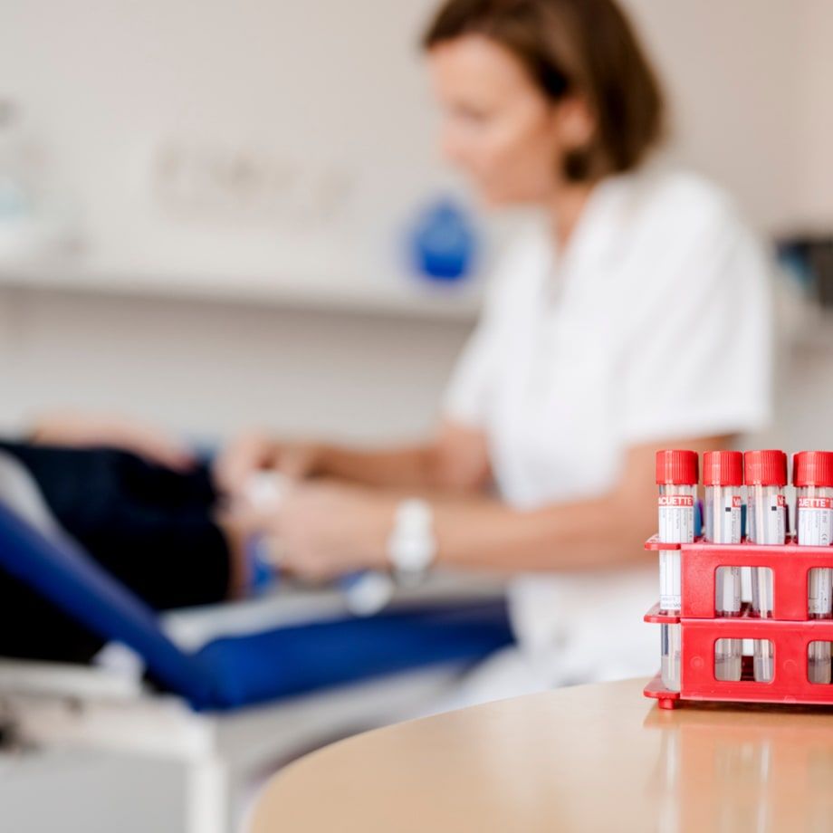 Im Vordergrund ein Ständer mit Blutentnahmeröhrchen mit roten Deckeln; im Hintergrund unscharf eine Pflegeperson bei einer Blutabnahme.